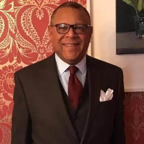 Patrick Swygert smiling and wearing a dark suit with a red tie in a warm tone, matching red-and-white pocket square and white shirt standing before a colorful red-and-gold wall and the edge of a golden framed portrait.
