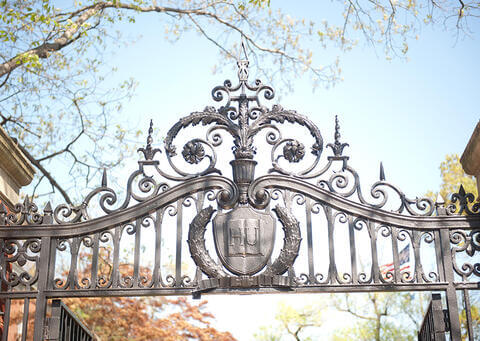 Iron-wrought Howard University Gate with decorative filligree and "HU" set at the center of a stylized book. Trees in autumn foliage and a blue sky are in the background.